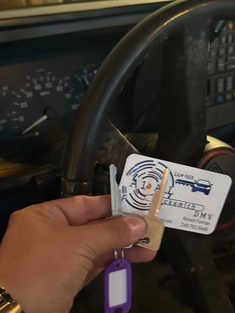 Two cut metal keys on a ring with a purple tag and an A1 All Locksmith DMV business card in front of an older vehicle’s steering wheel and analog gauges
