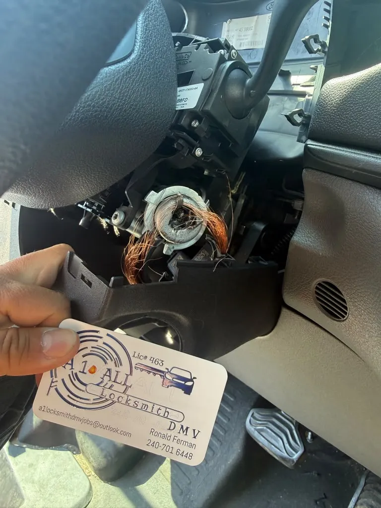Steering column with the lower cover removed, showing a damaged ignition lock housing and exposed wiring beside an A1 All Locksmith DMV business card