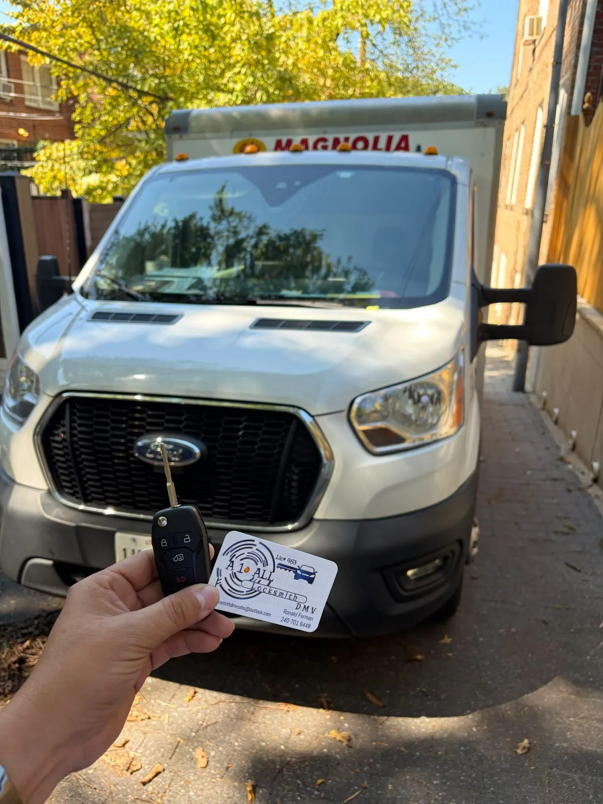 Hand holding a Ford flip remote key and an A1 All Locksmith DMV business card in front of a white Ford Transit box truck