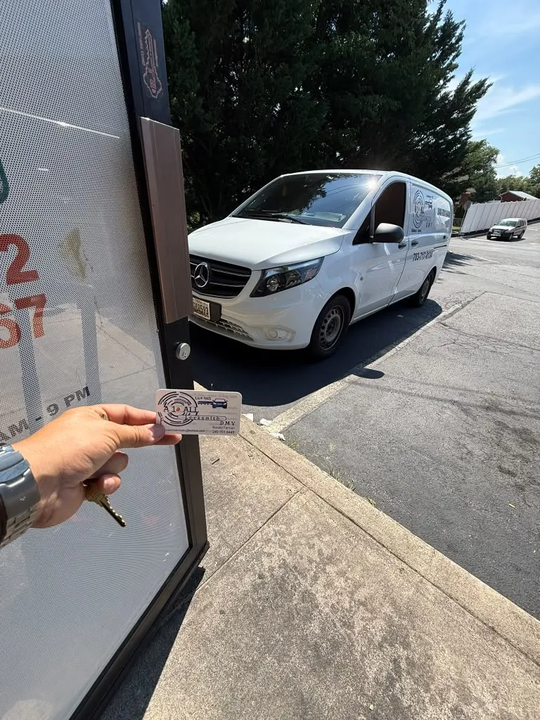 Hand holding a key and an A1 All Locksmith DMV business card at a glass storefront door, with a white van parked outside