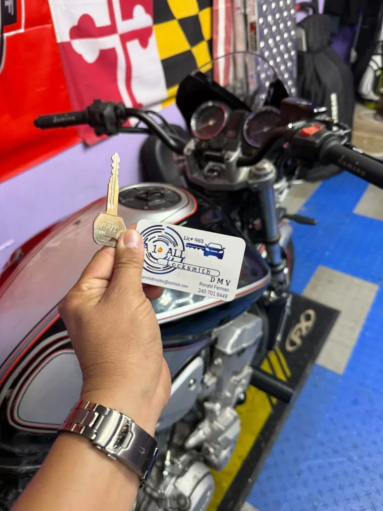 Cut motorcycle key and A1 All Locksmith DMV business card held above a motorcycle fuel tank in a garage with a Maryland flag on the wall