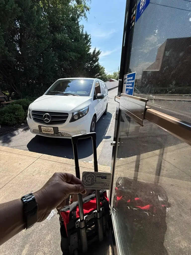 Hand holding an A1 All Locksmith DMV business card above a rolling tool bag at an open glass storefront door, with a white van parked at the curb